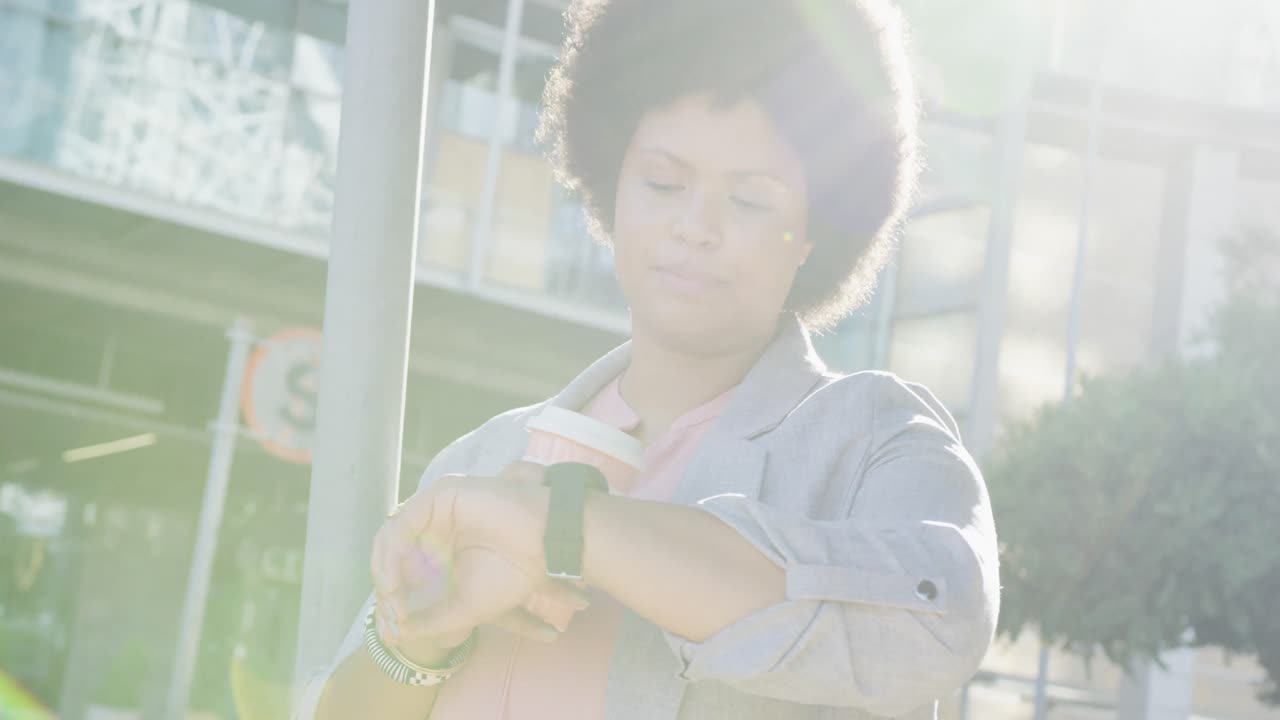 Plus size biracial woman checking smartwatch and drinking coffee in city