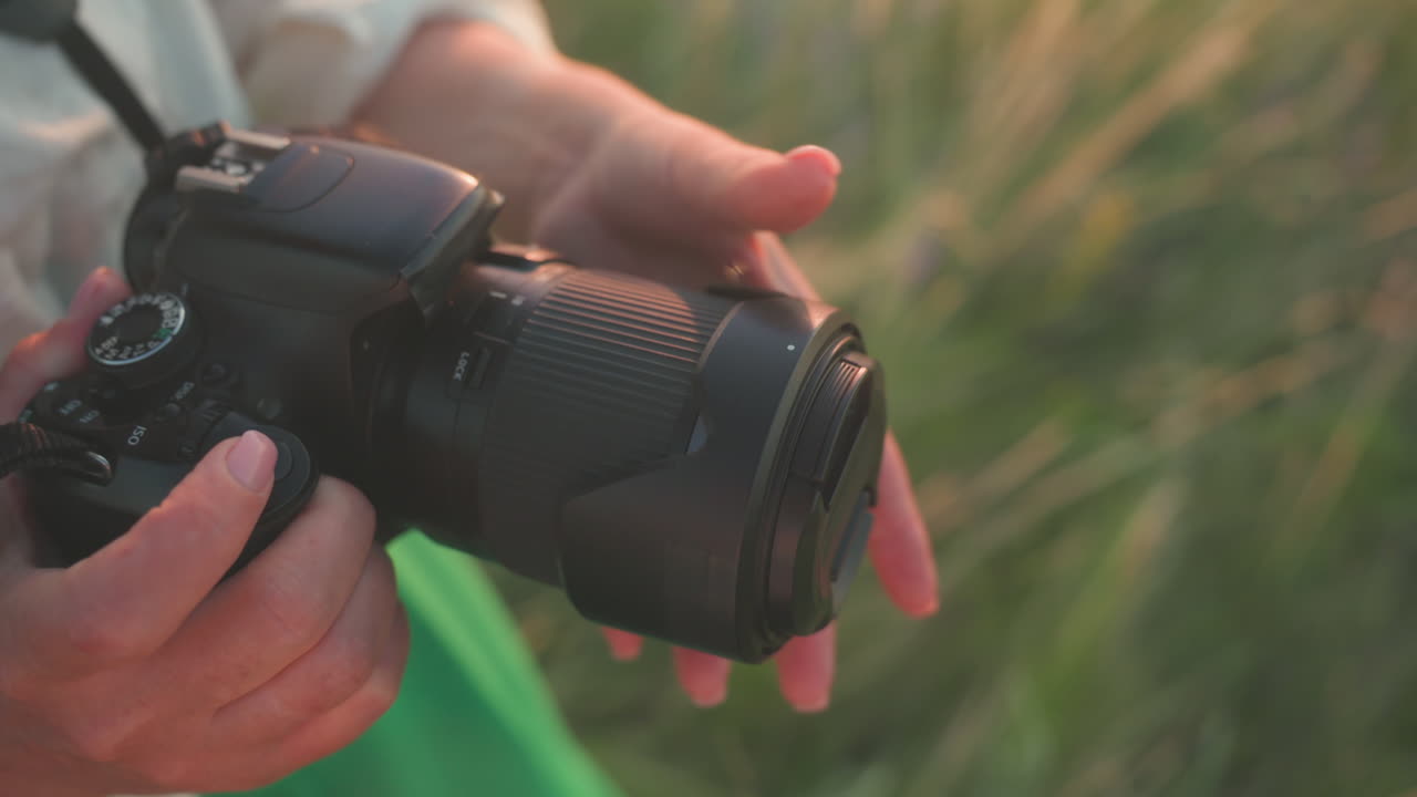 Close up partial view of lady hands gently closing lens cover on camera in warm golden light, camera strap around neck, standing in sunlit field with green dress visible