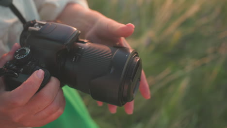 close up partial view of lady hands gently closing lens cover on camera in warm golden light, camera strap around neck, standing in sunlit field with green dress visible