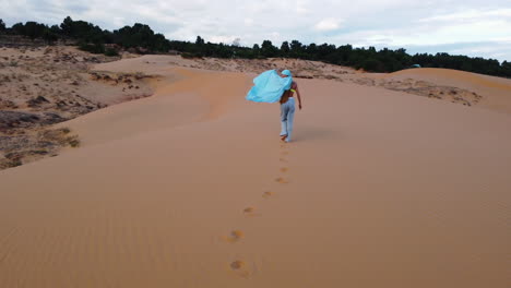 Aéreo,-Modelo-Con-Hijab-Azul-Caminando-Por-La-Arena-Del-Desierto,-Ondeando-Tela-De-Seda-En-El-Viento