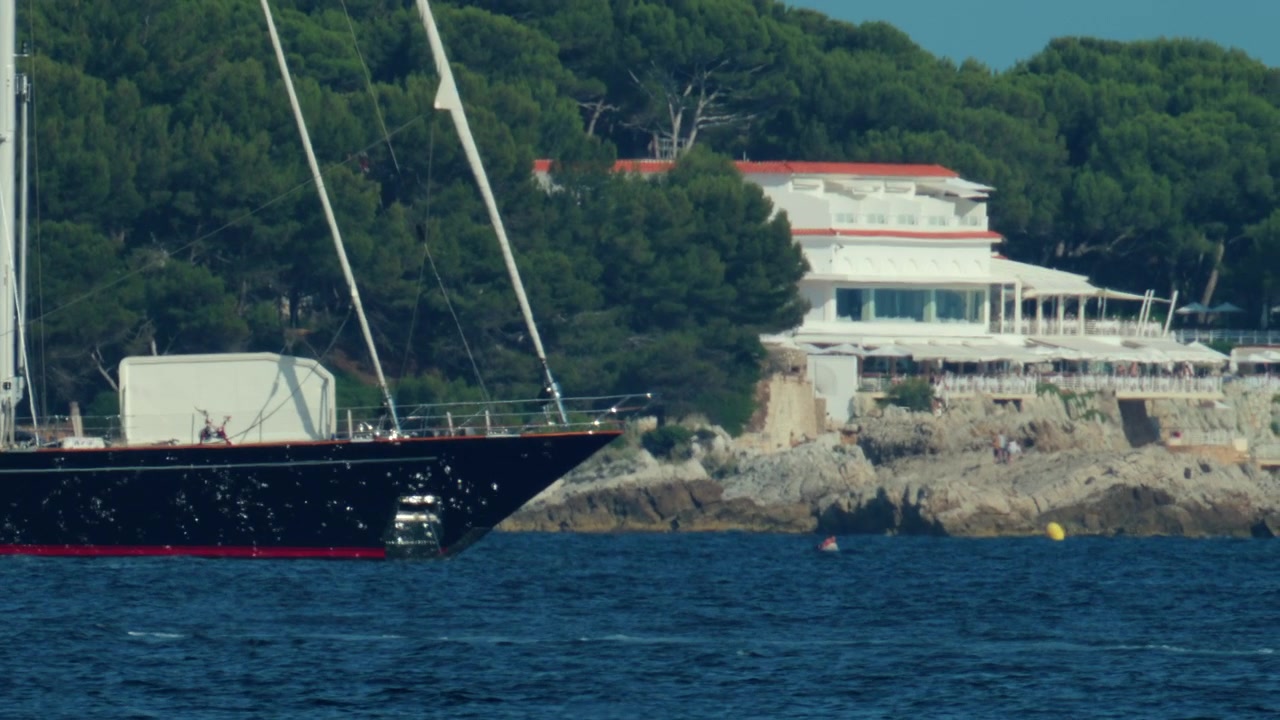 Yacht a vela con scafo nero che naviga su un mare blu con la costa mediterranea e alberi verdi sullo sfondo, a simboleggiare il viaggio di lusso.