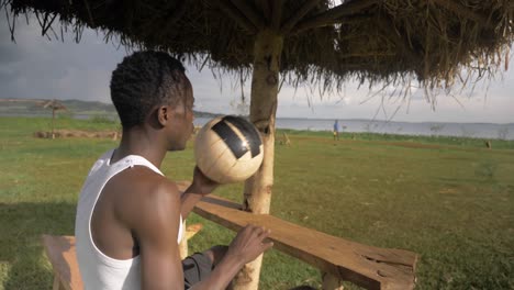 a close up shot of a young african man with a football relaxing under a beach hut on the shores of lake victoria