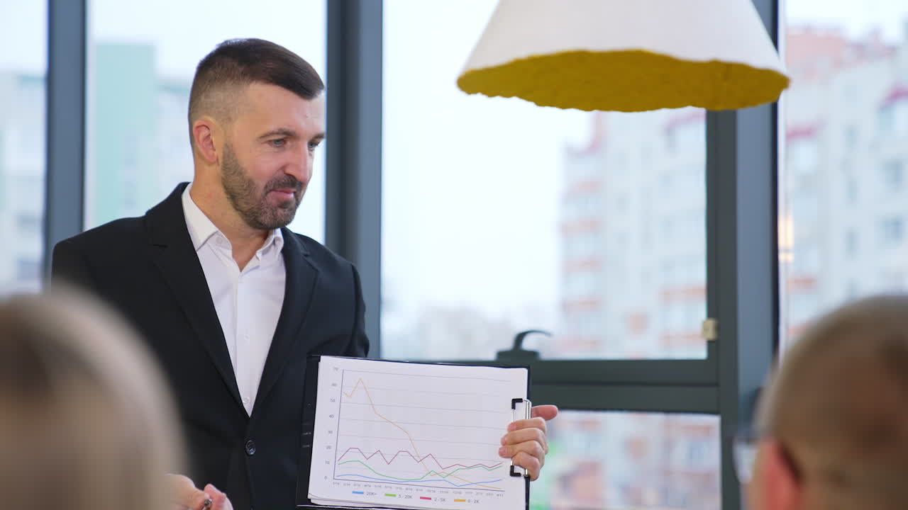 Bearded businessman wearing black suit holds a chart in his hands. Boss listens to his employees' ideas about the graphs.