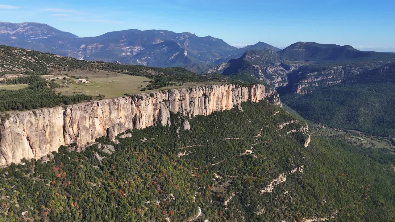 Aerial perspective of stunning cliffs surrounded by dense forests, mountains, and open fields under a bright blue sky in Catalonia.