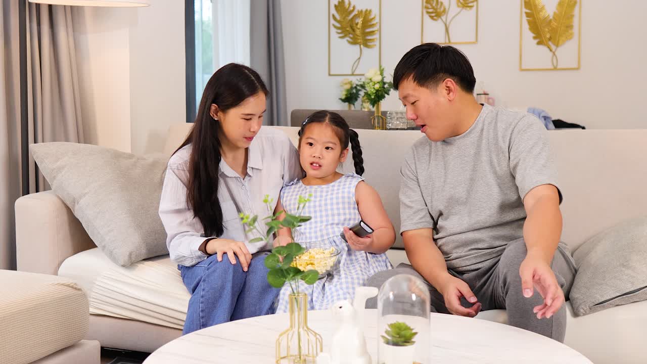Parents and daughter interact warmly on couch, exchanging remote, in bright, cozy home setting