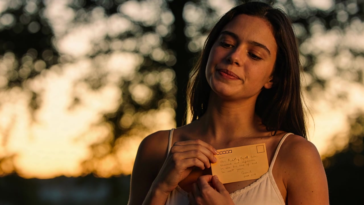 Donna con un top bianco che tiene in mano un biglietto scritto a mano giallo-arancio, legge e sorride sotto un bokeh al tramonto.