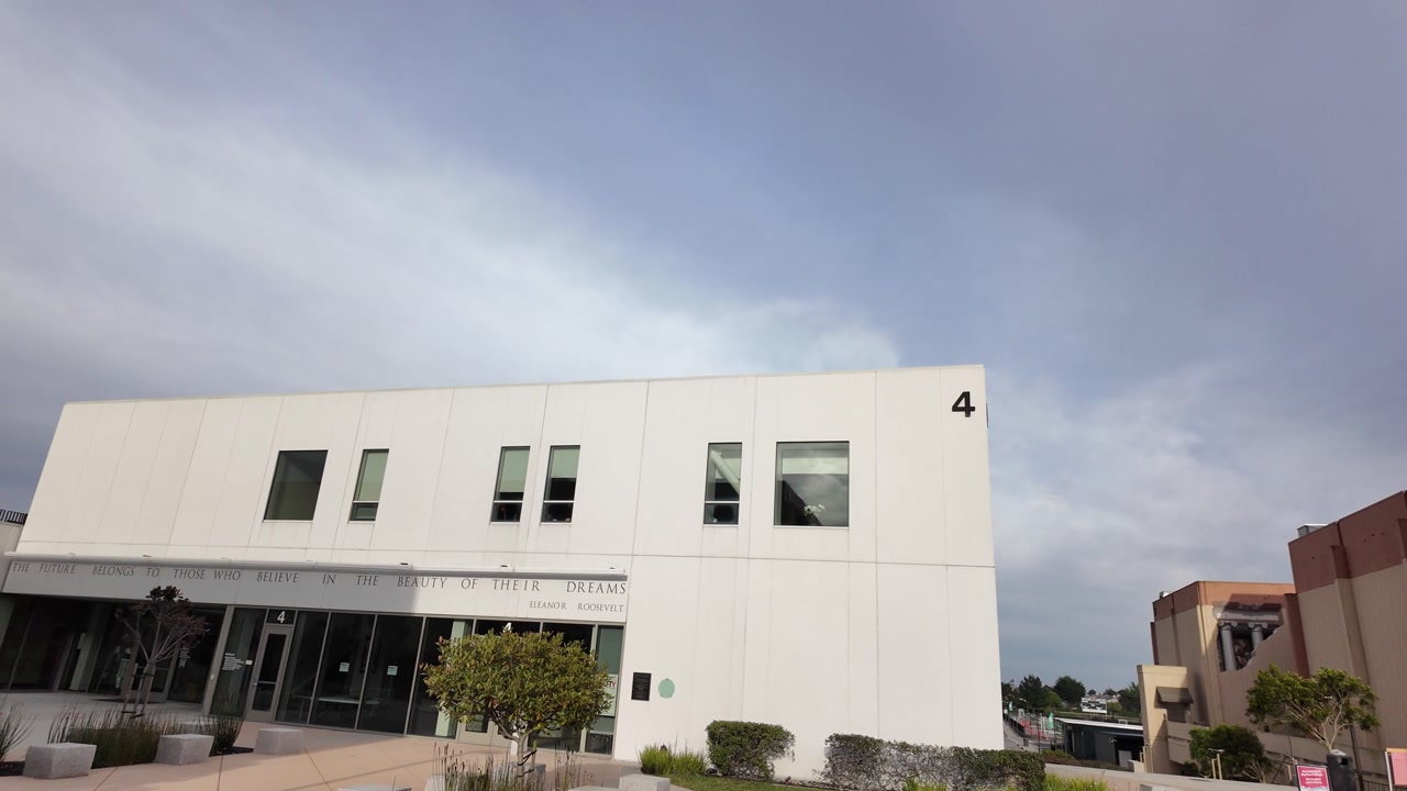 Wide-angle walking shot showing the minimalist white facade of a modern building on the Skyline College campus, located in San Bruno, California, under an overcast sky on a cloudy day
