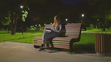 young girl sitting on park bench at night, deeply engaged with tablet under soft park lighting, surrounding scene shows people strolling in background