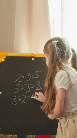 Girl writes task answer on blackboard while younger child shows plastic tag with digit at home. Elder sister teaches mathematics with boy sibling in class