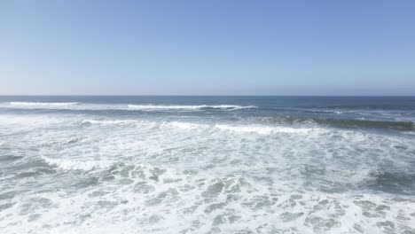 white foam and sand are churned as waves break, clear skies, rolling ocean, aerial