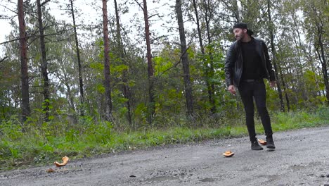 man destroying an orange halloween pumpkin in pieces by throwing it on the ground in slow motion during autumn with cloudy weather october. man wears black clothing and a leather jacket, has a cap on.
