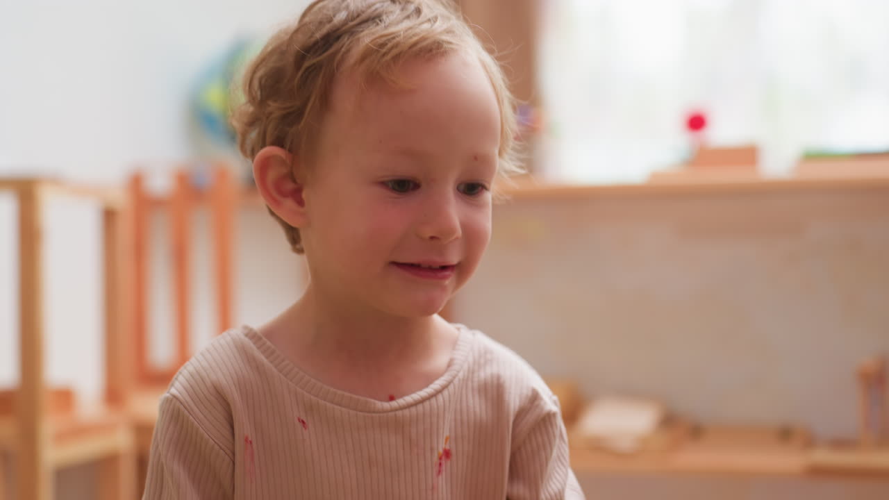 Fröhliches kleines Kind, das lächelt und aufgeregt in Richtung Hintergrund zeigt, während es an einer Aktivität im Klassenzimmer teilnimmt und dabei Neugier und Freude ausdrückt, umgeben von Holzmöbeln