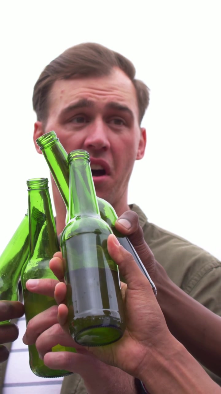 Vertikales Video: Grüne Flaschen klirren, Mann in olivfarbenem Hemd spricht, während Freunde im Garten anstoßen.