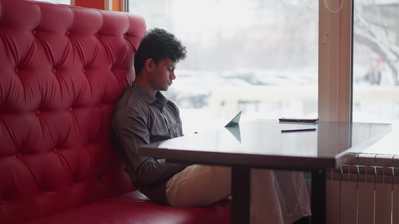 Young man sitting on red cushioned bench by window, holding paper and pen while looking thoughtfully at document, soft daylight and wintry outdoor scene contrast warm indoor setting, suggesting focus