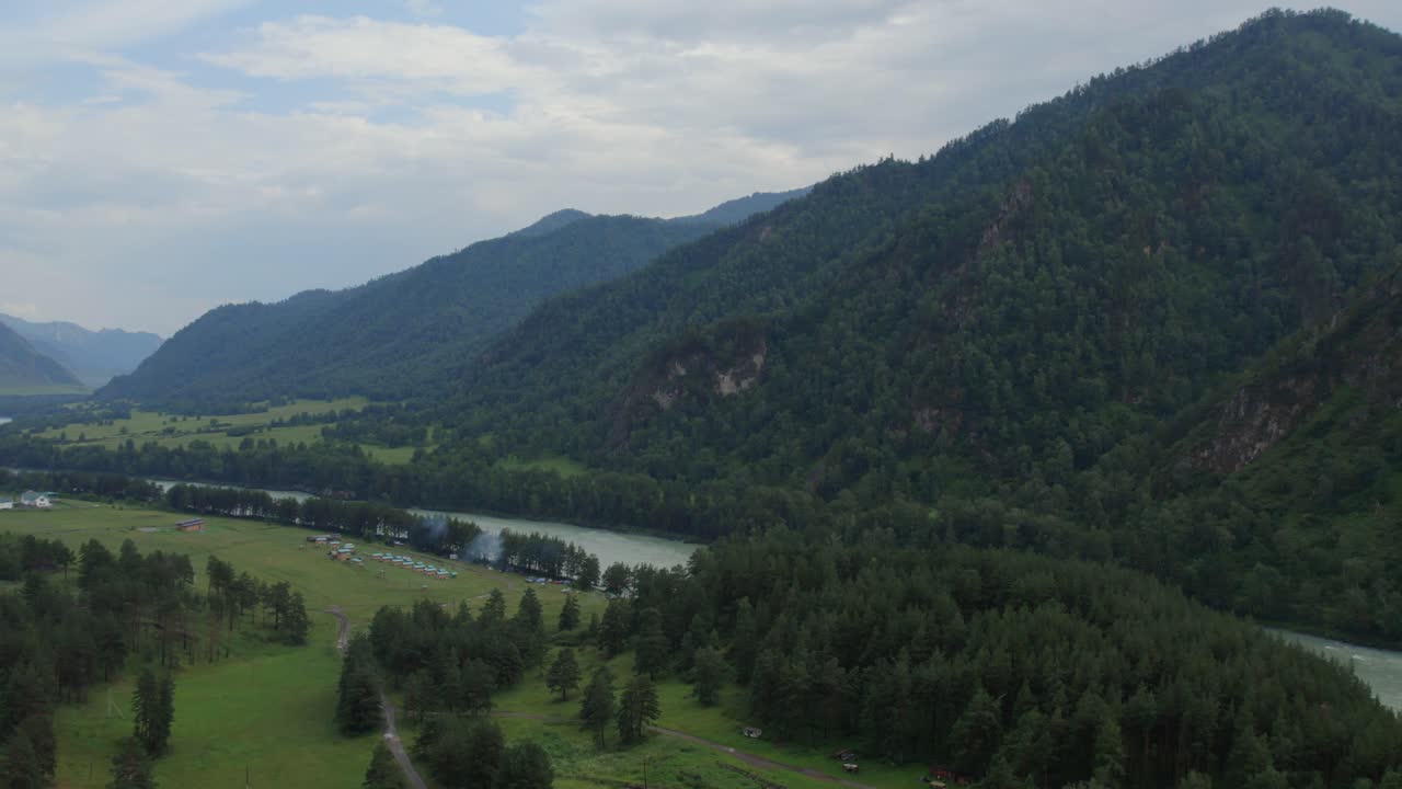 Drohnenaufnahme des Katun-Flusses, der sich durch ein grünes Tal mit Kiefernwald und Berghängen in der Altai-Republik, Russland, unter einem bewölkten Sommerhimmel schlängelt.