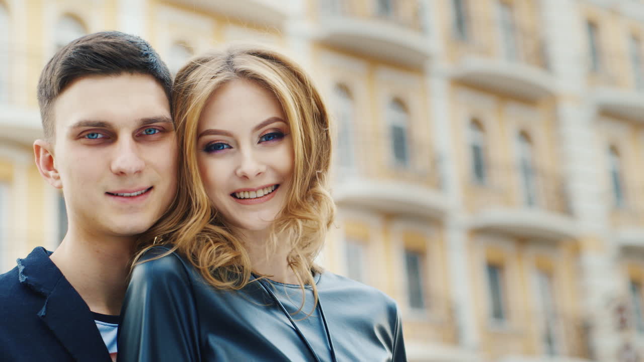 Uomo e donna sorridono guardando la telecamera. Sullo sfondo c'è un video leggermente sfocato di un edificio a più piani.