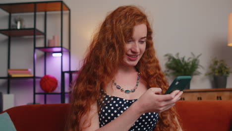 Young-redhead-girl-at-home-couch-use-smartphone-typing-browsing-say-wow-yes-found-out-great-big-win