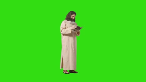 studio shot of man wearing robes and sandals with long hair and beard representing figure of jesus christ reading bible on green screen 1