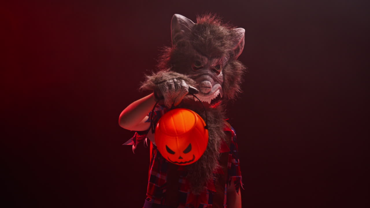 대인간 괴물 의상을 입은 아이의 스튜디오 은 은 연기 배경 조명과 함께 할로윈에 속임수 또는 대접 커 모양의 잭 o'lantern 버 2를 들고 있습니다.