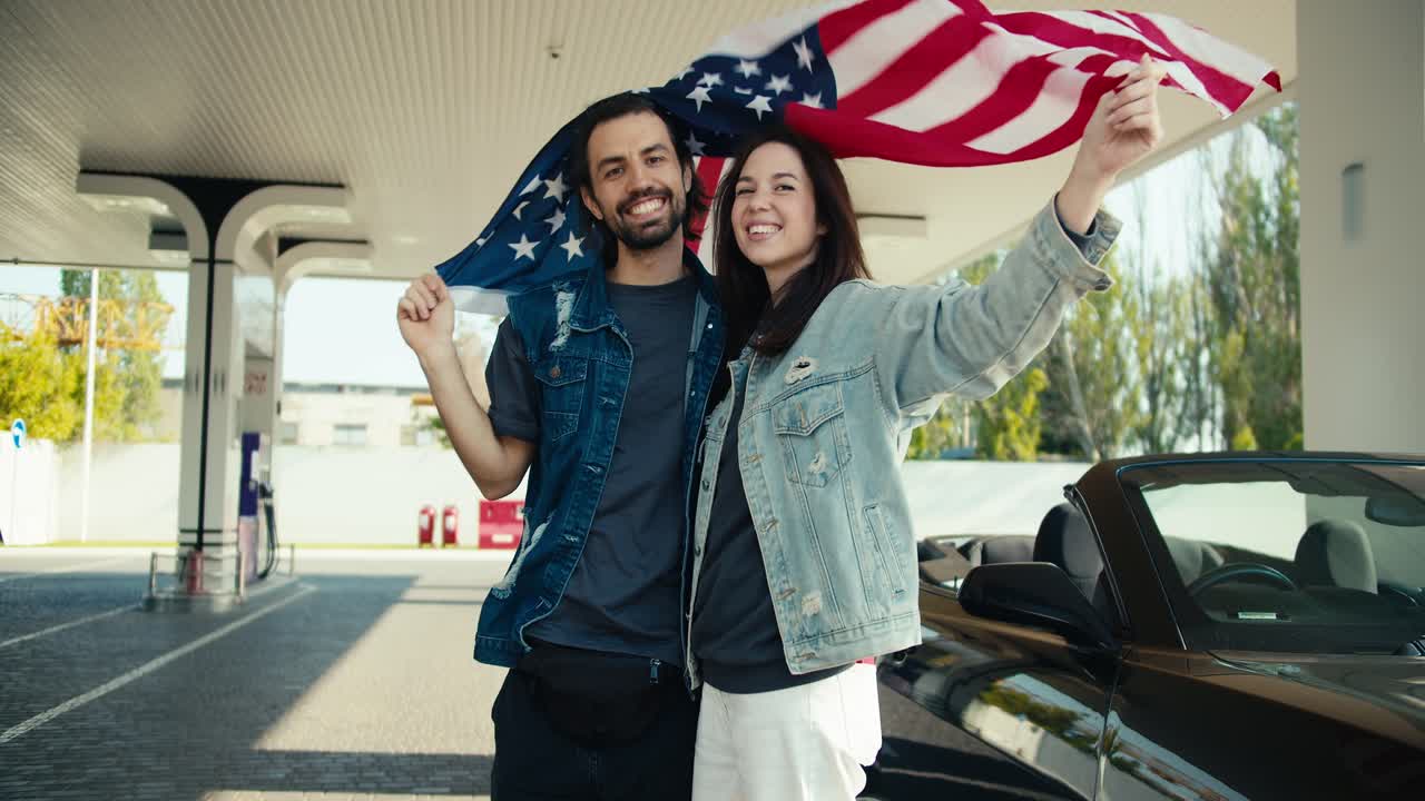 una pareja feliz de un chico y una chica morena en chaquetas de cuero levantan la bandera de los estados unidos sobre sus cabezas, se paran cerca de un gris