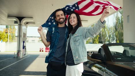 a happy couple of a guy and a brunette girl in leather jackets raise the us flag above their heads, they stand near a gray