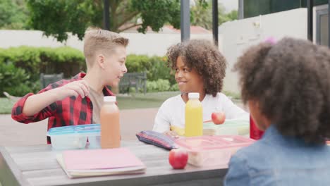 Video-of-three-diverse-schoolchildren-talking-at-lunch-sitting-in-schoolyard