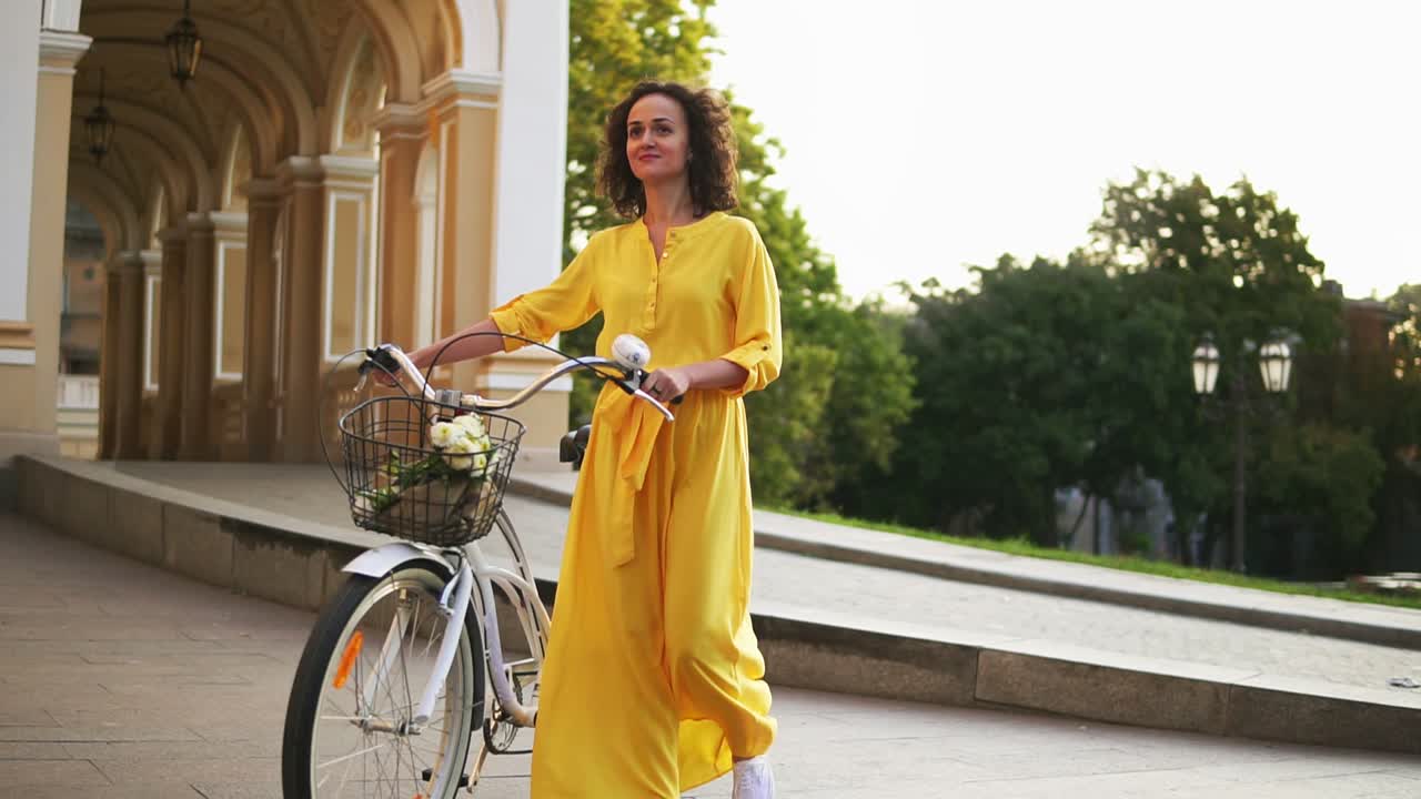 Smiling young girl in a long yellow dress walking holding her city bicycle handlebar with flowers in its basket in the city