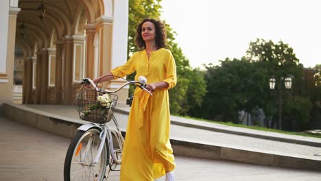 smiling young girl in a long yellow dress walking holding her city bicycle handlebar with flowers in its basket in the city