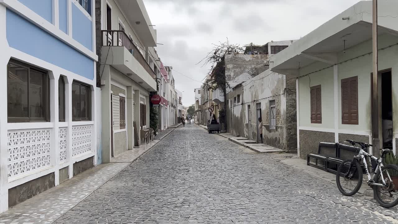 Zoom out shot of a cobblestone street in Sal Rei, Cape Verde, with blend of rustic and modern buildings. The tranquil urban atmosphere features local charm, bicycles, and a peaceful street scene