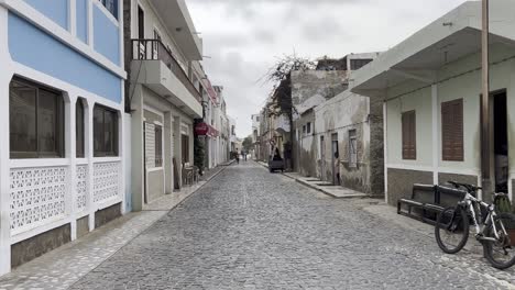 zoom out shot of a cobblestone street in sal rei, cape verde, with blend of rustic and modern buildings. the tranquil urban atmosphere features local charm, bicycles, and a peaceful street scene