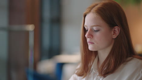 thoughtful brooding remote working red haired woman sitting infront of a laptop or notebook in casual outfit on her work desk in her modern airy bright living room home office with many windows