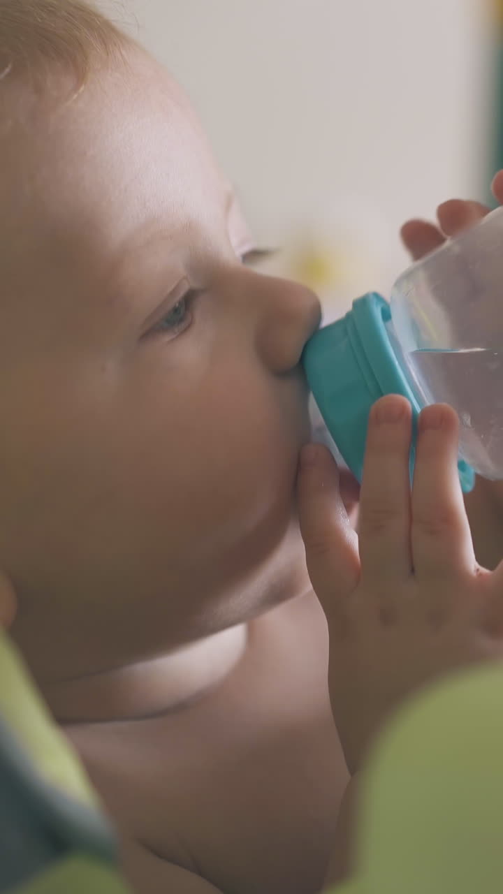 funny little child drinks water from bottle with sister help sitting in green highchair in light room closeup