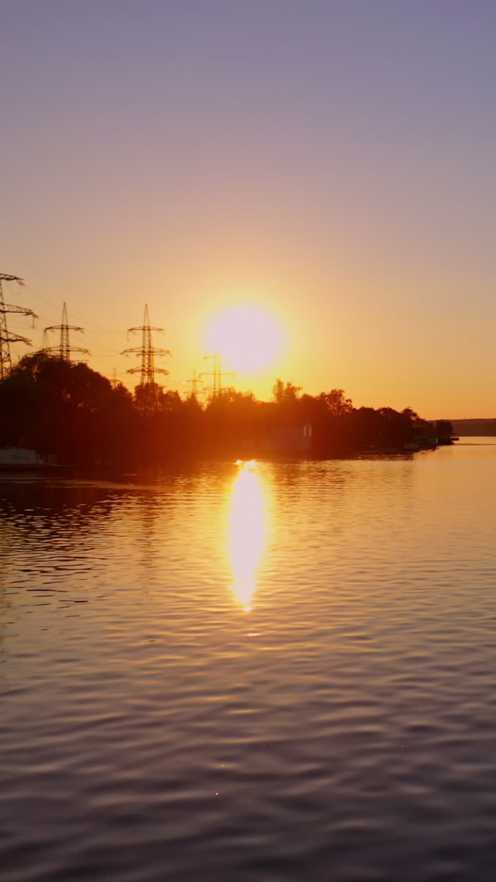 Evening river against the transmission lines at sunset. Flight over the river on the background of dark silhouettes of high voltage electric line. Aerial view. Vertical video