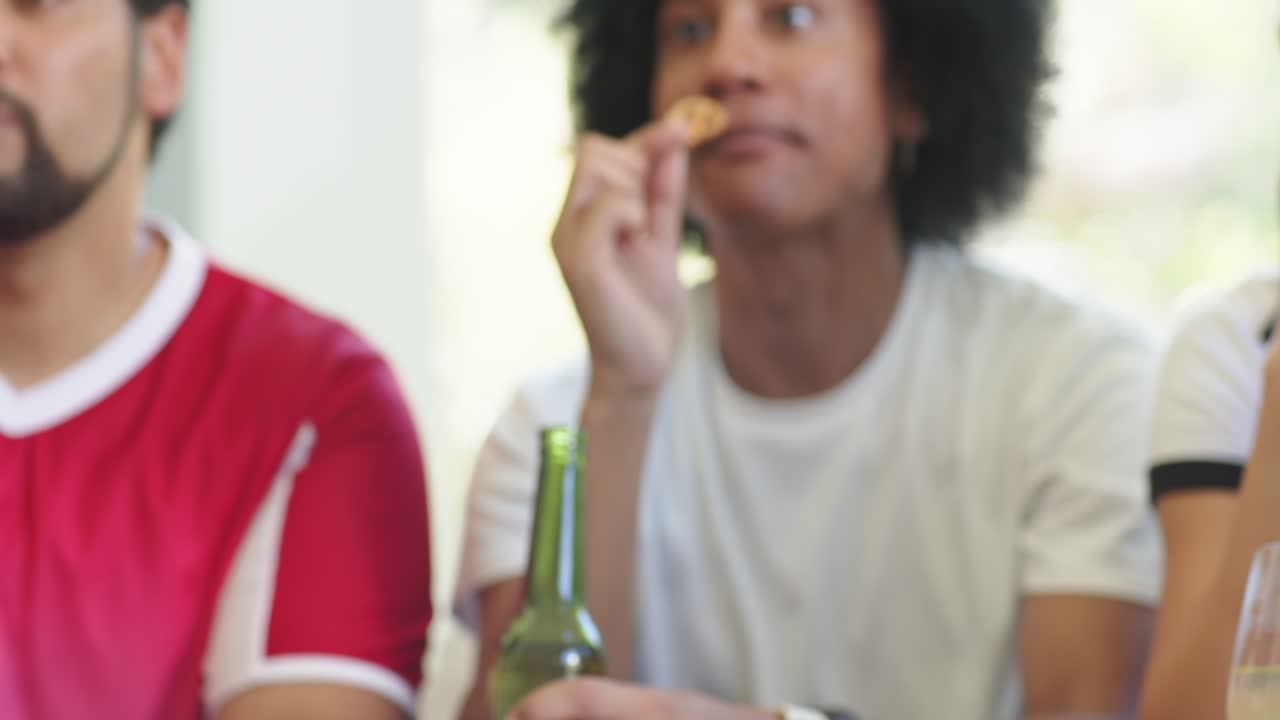 Diverse men grabbing snack on couch, causing curly man eating pretzel reacting to TV, green bottle