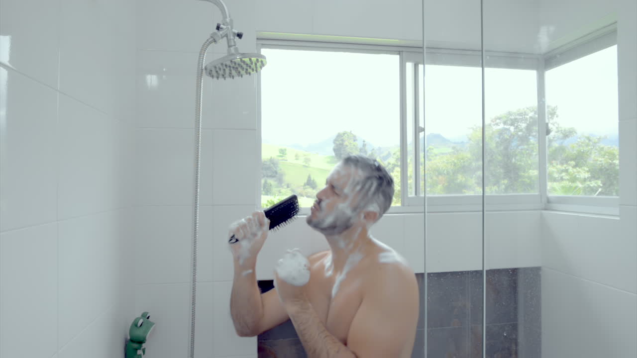 Muž zpívá a tančí ve sprše a nanáší si šampon na vlasy.