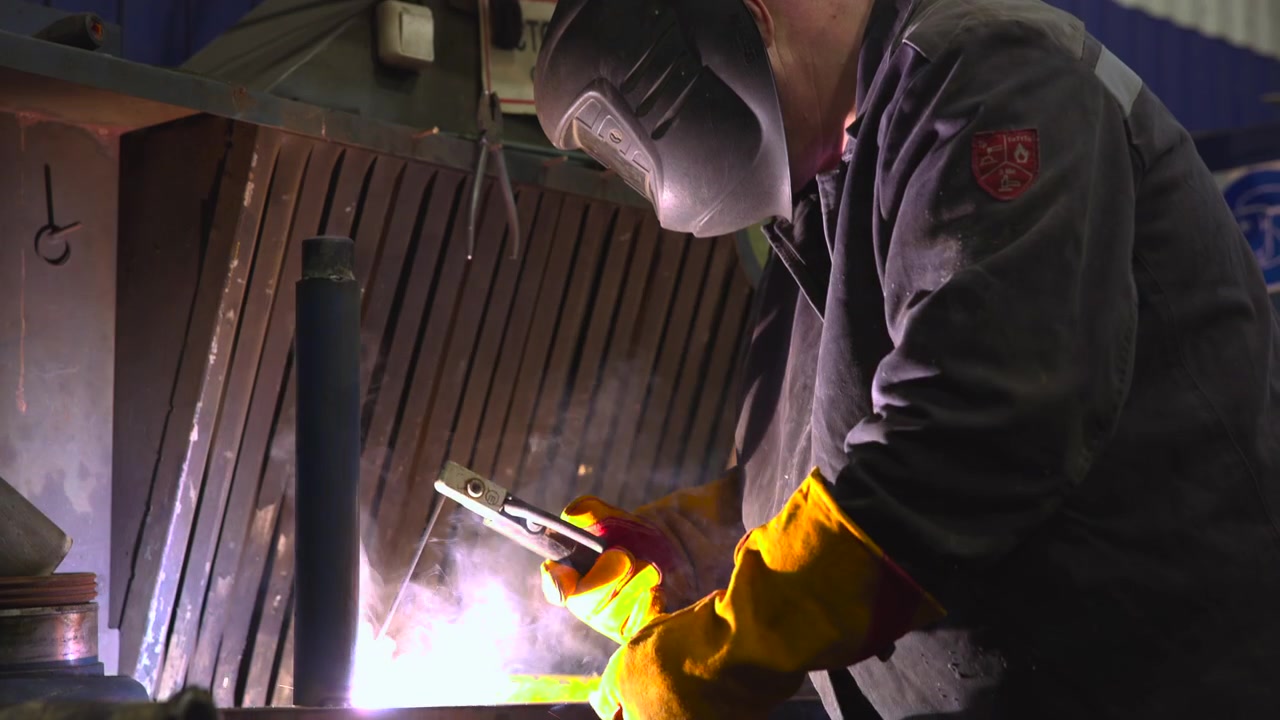 Close medium view of welder leaning over metal bench while holding electrode clamp with both gloved hands as bright welding arc emits white-magenta light, smoke, and sparks across industrial workshop