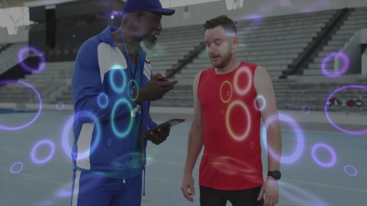 Allenatore che parla con un atleta su una pista di tecnologia sportiva, mostrando dati tramite un tablet con anelli al neon.