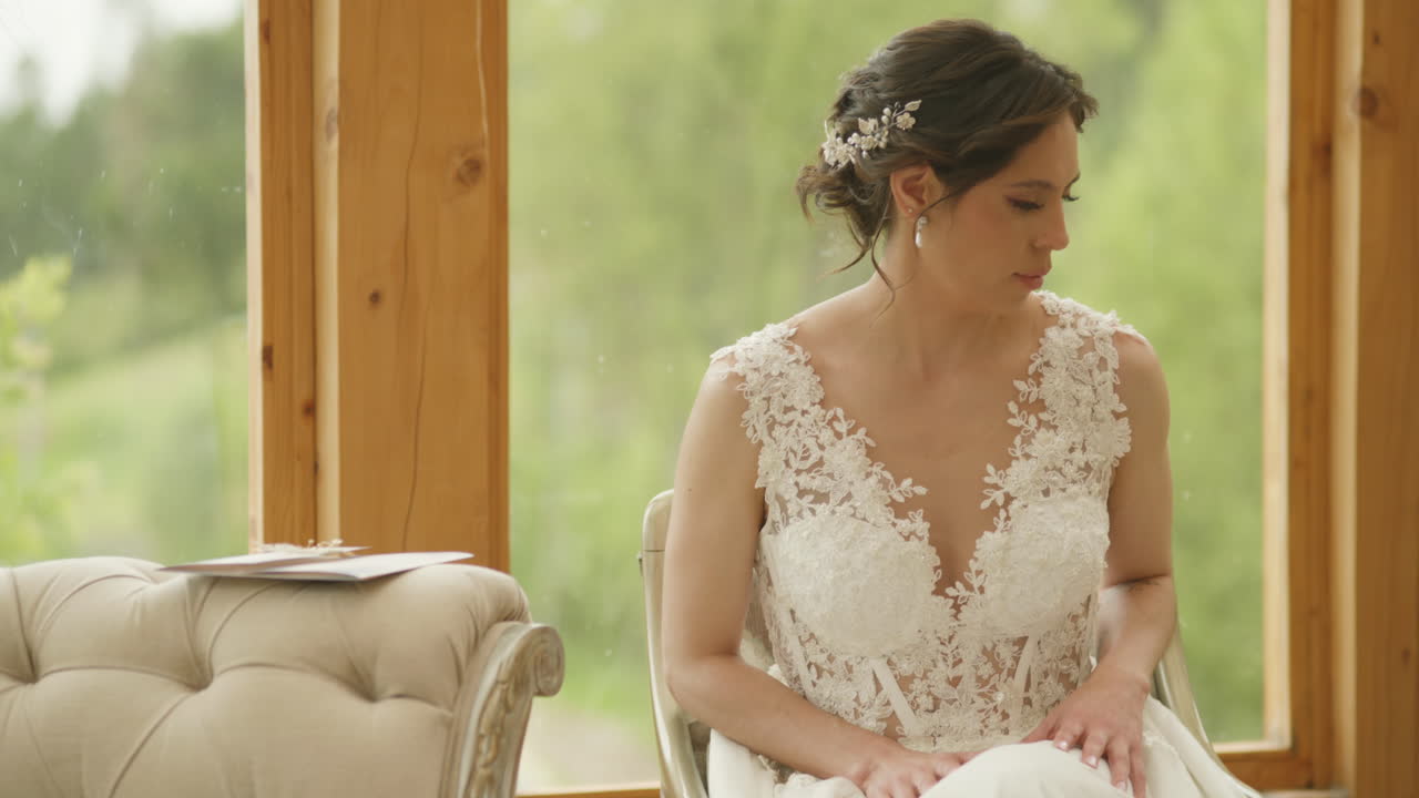 A bride sits on a sofa, calmly gazing out the window overlooking the countryside. The wedding invitation rests beside her, in an intimate, elegant, and thoughtful scene.