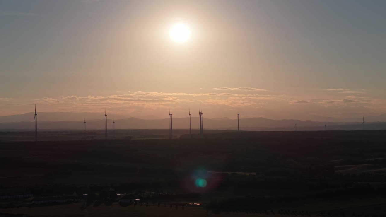 Silhouettes of wind turbines standing tall against the setting sun over a wide landscape. The scene highlights renewable energy production in harmony with nature and the evening sky