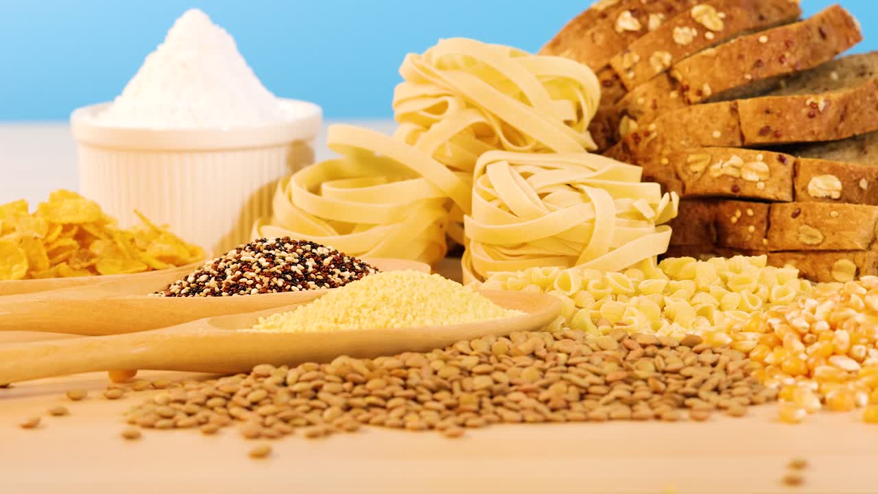A variety of grains, pasta, and bread arranged on a wooden surface with a blue background, under bright lighting