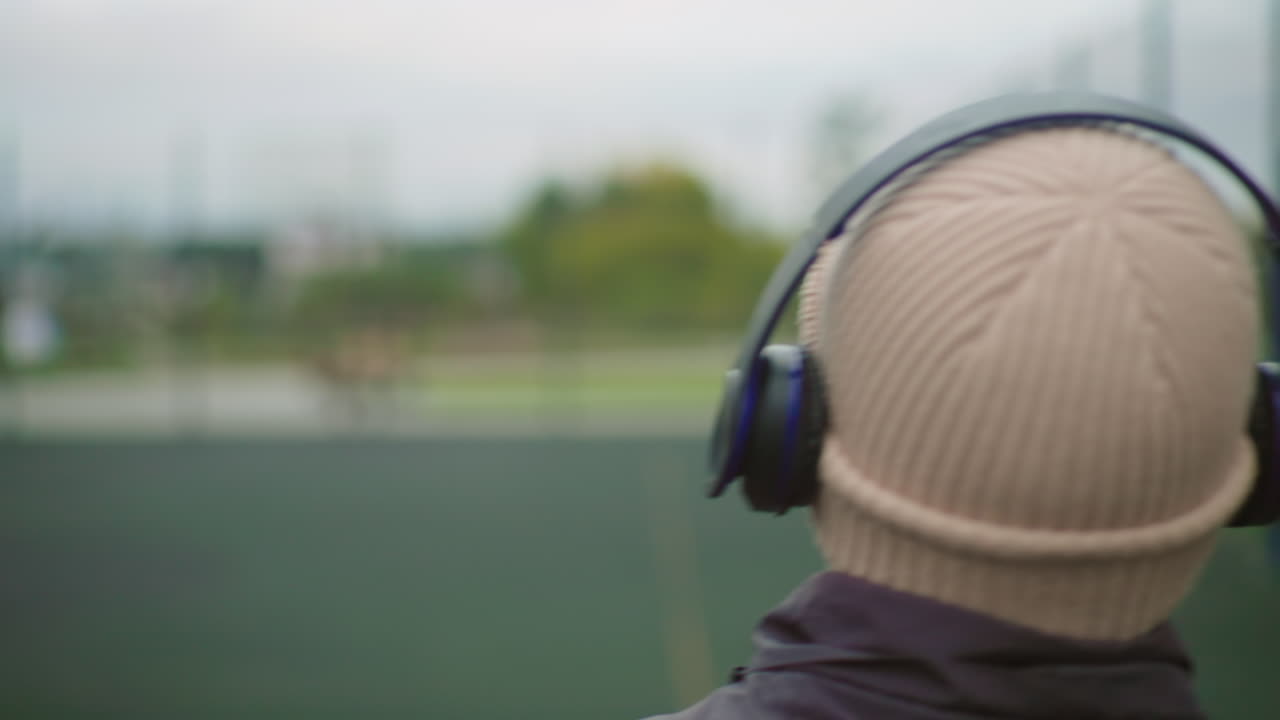 Persona con cuffie e berretto rivolta verso un campo, di spalle alla camera in un parco urbano, campo sportivo sfocato sullo sfondo, luce fredda e nuvolosa, giacca di lana e cuffie wireless che indicano l'ascolto di musica, atmosfera
