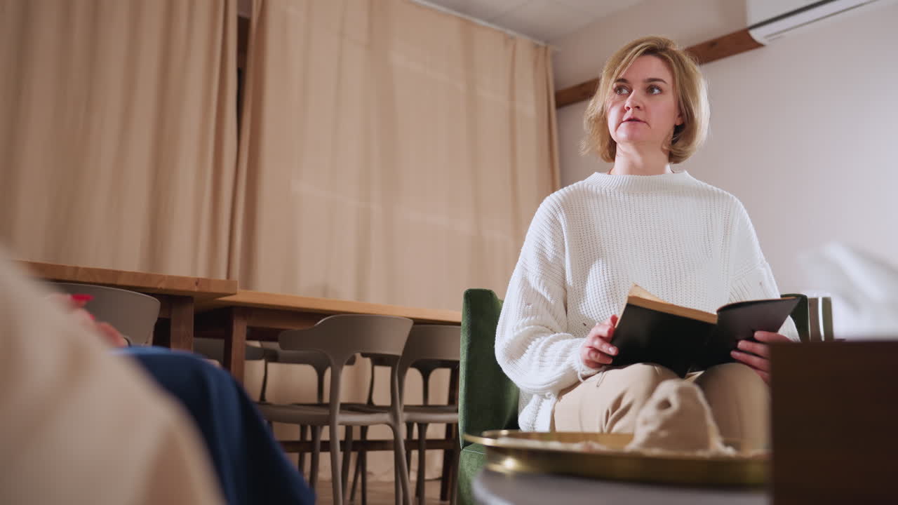 Conversation partner sits calmly holding book during dialogue in cozy therapy lounge, engaging in shared reading moment with blurred listener in foreground and warm ambient room setting