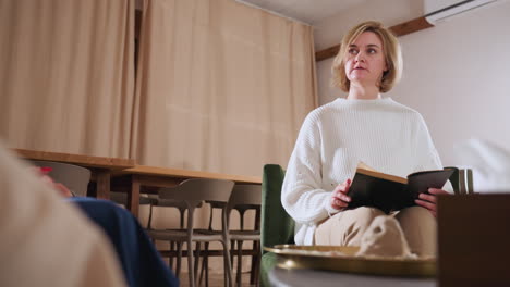 conversation partner sits calmly holding book during dialogue in cozy therapy lounge, engaging in shared reading moment with blurred listener in foreground and warm ambient room setting