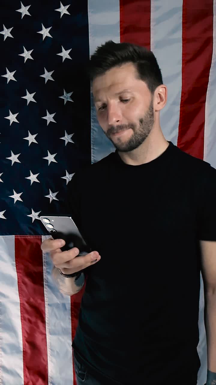 Tattooed man with short hair and a beard standing in front of a vertical American flag and scrolling his phone with a smile and while nodding. Person chatting on the phone, looks like a patriot