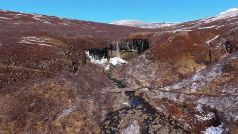 Fondo-De-Montaña-En-El-Horizonte-En-Las-Cascadas-Islandesas-De-Svartifoss-Zoom-Aéreo-De-Drones-En-Agua-En-Cascada,-Paisaje-Negro-Oculto-Del-Parque-Nacional