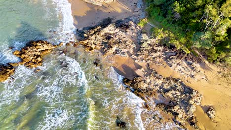 drone footage captures waves hitting rocks on a sunlit beach with lush greenery