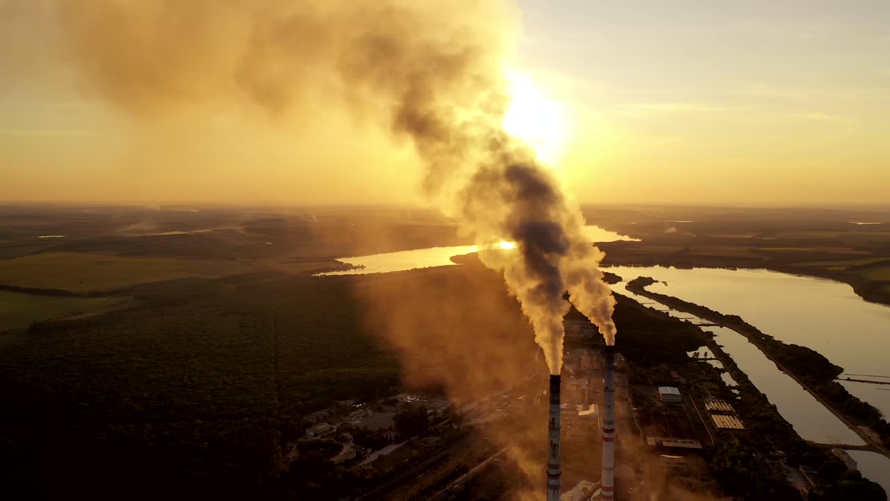 Industrial pipes with smoke in nature. Metallurgical plant among green fields at beautiful sunset. Aerial view. Camera moves right.