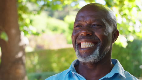 Portrait-Of-Smiling-Senior-Man-Standing-Outdoors-In-Garden-Park-Or-Countryside