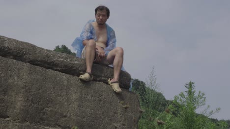푸른 플라스틱 레인코트를 입은 남자가 야외의 큰 바위 위에 앉아서 멀리 바라보고 있습니다.