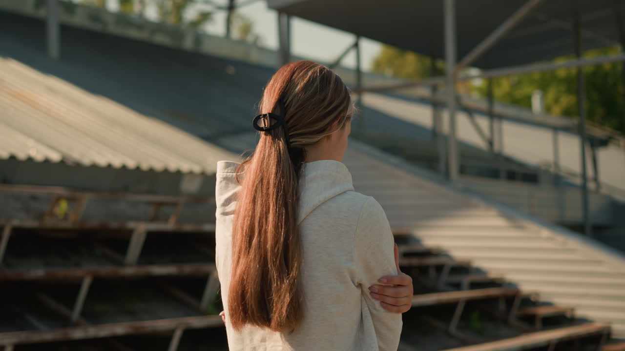 은 날에 빈 경기장 좌석 근처에서 걸어가는 긴 머리카락을 은 후두를 입은 여자의 뒷면, 손을 접고, 사려 깊고 반사적으로 보이며, 빈 관중석과 따뜻한 빛으로 둘러싸여 있습니다.
